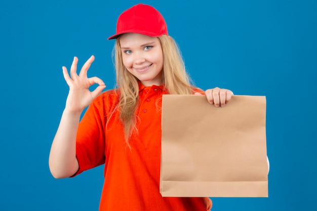 Entrega joven mujer vistiendo polo naranja y gorra roja sosteniendo el paquete de papel haciendo bien firmar sonriendo amable sobre fondo azul aislado