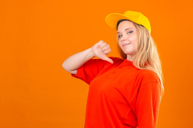 Entrega joven mujer vistiendo polo rojo y gorra amarilla mirando confiado apuntando a sí misma sonriendo sobre fondo naranja aislado