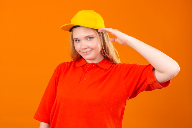 Entrega joven mujer vistiendo polo rojo y gorra amarilla saludando mirando confiado sobre fondo naranja aislado