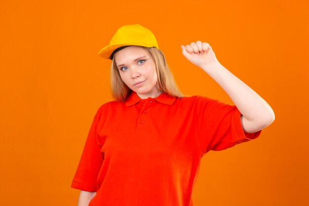 Entrega joven mujer vistiendo polo rojo y gorra amarilla sonriendo amable de pie con el concepto ganador de puño levantado sobre fondo naranja aislado