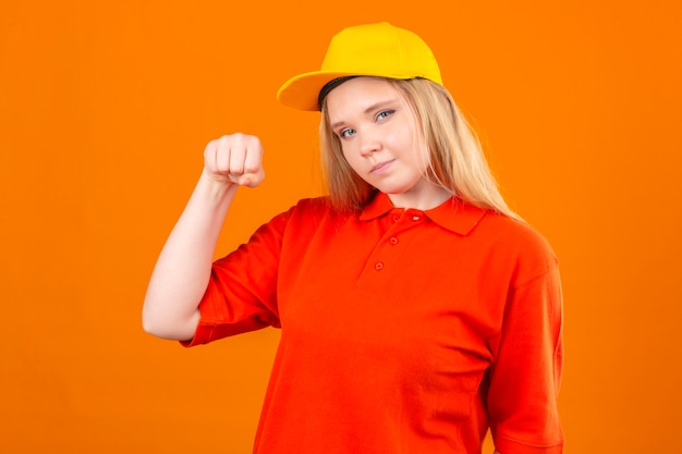 Entrega joven mujer vistiendo polo rojo y gorra amarilla sonriendo amable de pie con el concepto ganador de puño levantado sobre fondo naranja aislado