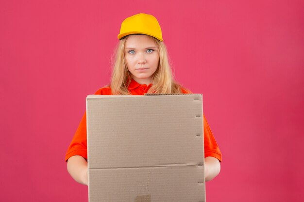 Entrega joven mujer vistiendo polo rojo y gorra amarilla sosteniendo una caja de cartón mirando a la cámara con cara seria sobre fondo rosa aislado