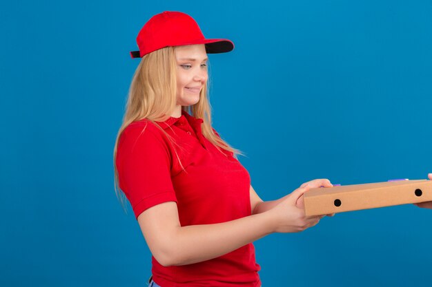 Entrega joven mujer vistiendo polo rojo y gorra dando caja de pizza a un cliente sonriendo amable sobre fondo azul aislado
