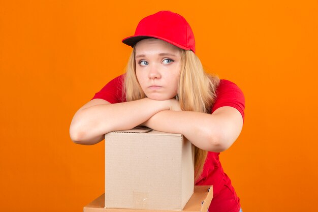 Entrega joven mujer vistiendo polo rojo y gorra esperando con la mano cruzada en la pila de cajas de cartón con aspecto cansado y enfermo sobre fondo naranja aislado