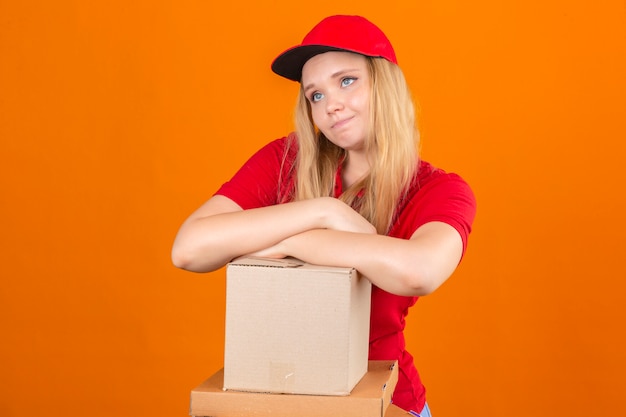 Foto gratuita entrega joven mujer vistiendo polo rojo y gorra esperando con la mano cruzada en la pila de cajas de cartón con aspecto cansado y enfermo sobre fondo naranja aislado