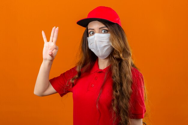 Entrega joven mujer vistiendo polo rojo y gorra en máscara protectora médica mirando sorprendido haciendo bien firmar sobre fondo naranja aislado