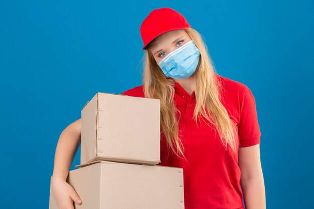 Entrega joven mujer vistiendo polo rojo y gorra en máscara protectora médica de pie con cajas de cartón con aspecto cansado y con exceso de trabajo sobre fondo azul aislado