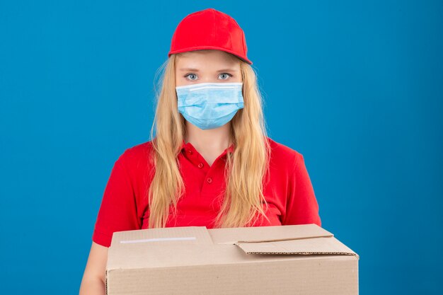 Entrega joven mujer vistiendo polo rojo y gorra en máscara protectora médica de pie con cajas de cartón mirando a cámara con cara seria sobre fondo azul aislado