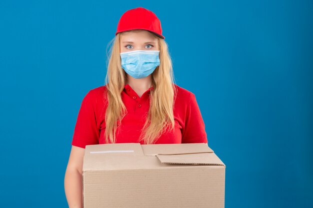 Entrega joven mujer vistiendo polo rojo y gorra en máscara protectora médica de pie con cajas de cartón mirando a cámara con cara seria sobre fondo azul aislado