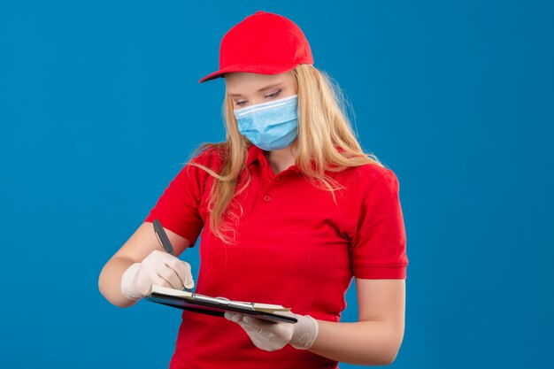 Foto gratuita entrega joven mujer vistiendo polo rojo y gorra en máscara protectora médica de pie con portapapeles escribiendo mirando seriamente sobre fondo azul aislado