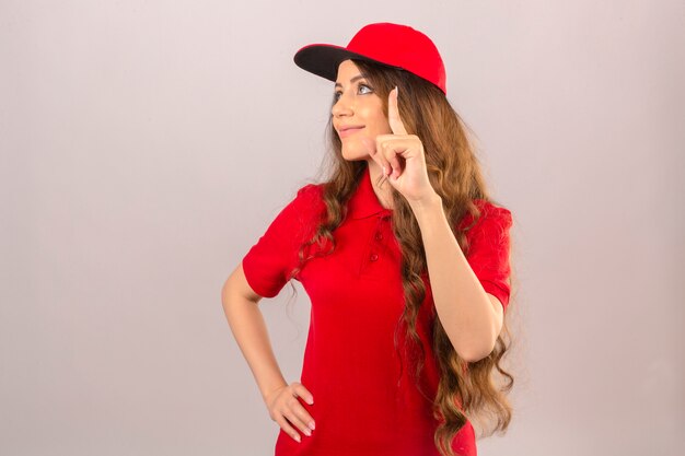 Entrega joven mujer vistiendo polo rojo y gorra mirando hacia arriba y apuntando con el dedo sonriendo sobre fondo blanco aislado