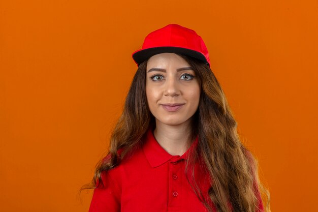 Entrega joven mujer vistiendo polo rojo y gorra con pelo rizado mirando a la cámara con sonrisa de confianza sobre fondo naranja aislado