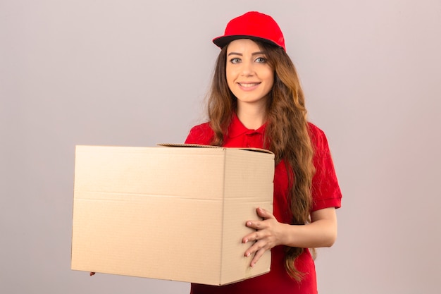 Entrega joven mujer vistiendo polo rojo y gorra de pie con caja de cartón sonriendo alegremente sobre fondo blanco aislado