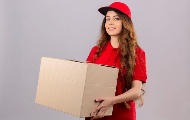 Entrega joven mujer vistiendo polo rojo y gorra de pie con caja de cartón sonriendo alegremente sobre fondo blanco aislado