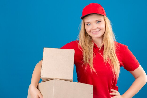 Entrega joven mujer vistiendo polo rojo y gorra de pie con cajas de cartón mirando a la cámara sonriendo amable sobre fondo azul aislado