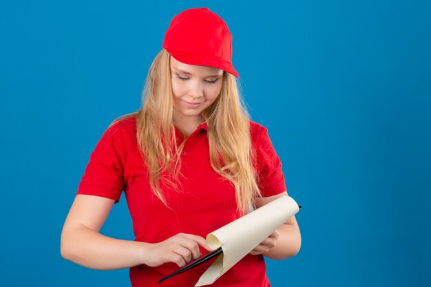 Entrega joven mujer vistiendo polo rojo y gorra de pie con portapapeles mirando espacios en blanco con una sonrisa en la cara sobre fondo azul aislado