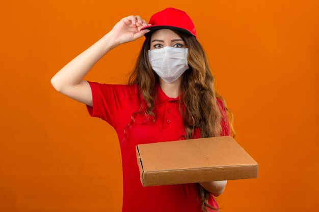 Entrega joven mujer vistiendo polo rojo en máscara protectora médica poniéndose la tapa de pie con pila de cajas de pizza mirando confiado sobre fondo naranja aislado