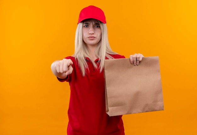 Entrega joven vistiendo camiseta roja y gorra sosteniendo el bolsillo de papel mostrando su gesto en la pared naranja aislada