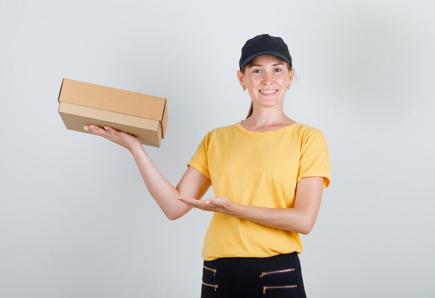 Entrega mujer mostrando caja de cartón en camiseta, pantalón, gorra y mirando contento