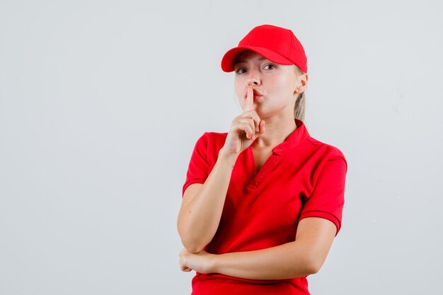 Entrega mujer mostrando gesto de silencio en camiseta roja y gorra y mirando con cuidado