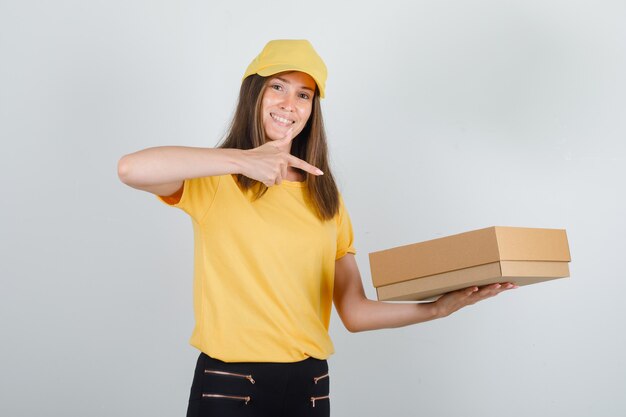 Entrega mujer señalando con el dedo a la caja de cartón en camiseta, pantalones, gorra y mirando alegre