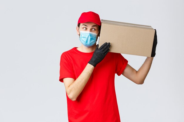 Entrega de paquetes y encomiendas, entrega en cuarentena covid-19, órdenes de transferencia. Mensajero curioso en uniforme rojo, guantes y mascarilla protectora, entregar la caja al cliente, traer el pedido sin contacto