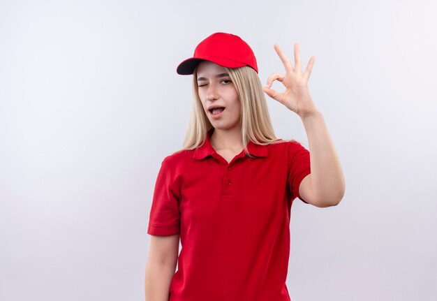 Entrega parpadeó joven vistiendo camiseta roja y gorra mostrando gesto okey en pared blanca aislada