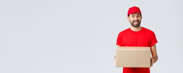 Foto gratuita entrega de pedidos, compras en línea y concepto de envío de paquetes. mensajero amable y sonriente con gorra de uniforme rojo y camiseta, repartiendo paquetes para los clientes. el empleado trae caja de paquetería, fondo gris.