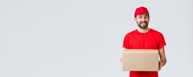 Entrega de pedidos, compras en línea y concepto de envío de paquetes. Mensajero amable y sonriente con gorra de uniforme rojo y camiseta, repartiendo paquetes para los clientes. El empleado trae caja de paquetería, fondo gris.