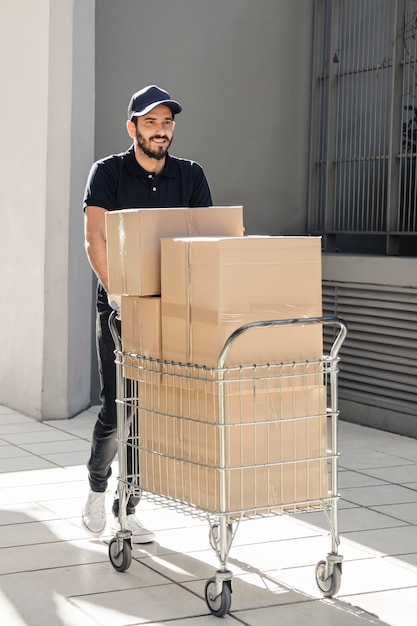 Entrega sonriente caminando sobre el pavimento con la carretilla llena de cajas de cartón