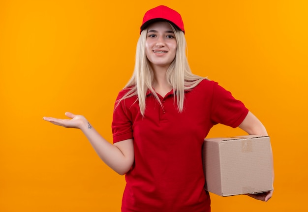 Entrega sonriente joven vestida con camiseta roja y gorra en la caja de sujeción de corsé dental y puntos al lado sobre fondo naranja aislado