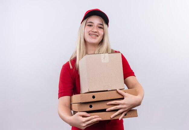 Entrega sonriente joven vestida con camiseta roja y gorra en caja de sujeción dental y caja de pizza sobre fondo blanco aislado