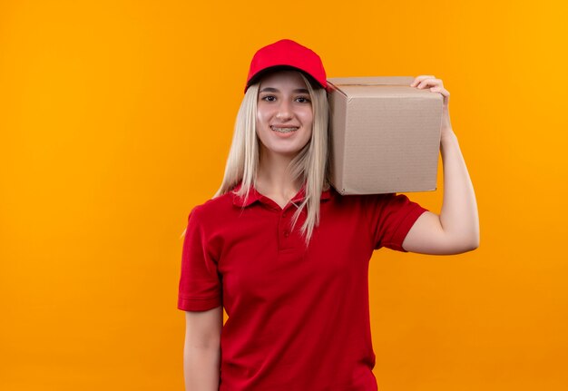 Entrega sonriente joven vestida con camiseta roja y gorra en caja de sujeción de soporte dental en el hombro sobre fondo naranja aislado