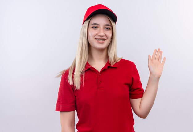 Entrega sonriente joven vestida con camiseta roja y gorra en corsé dental mostrando gesto de saludo sobre fondo blanco aislado