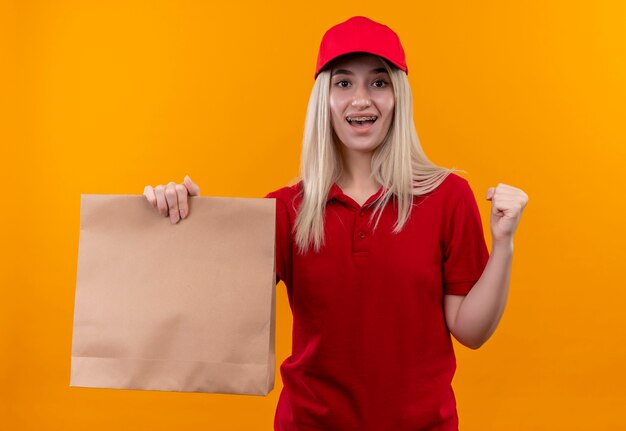 Entrega sonriente joven vestida con camiseta roja y gorra en corsé dental sosteniendo el bolsillo de papel mostrando sí gesto sobre fondo naranja aislado