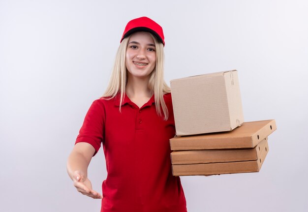 Entrega sonriente joven vestida con camiseta roja y gorra en corsé dental sosteniendo la caja de pizza sosteniendo la mano a la cámara sobre fondo blanco aislado