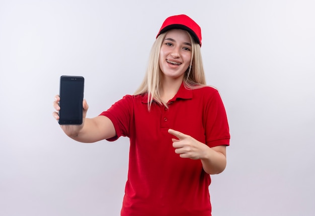 Entrega sonriente joven vestida con camiseta roja y gorra en puntos de apoyo dental en el teléfono en la mano sobre fondo blanco aislado