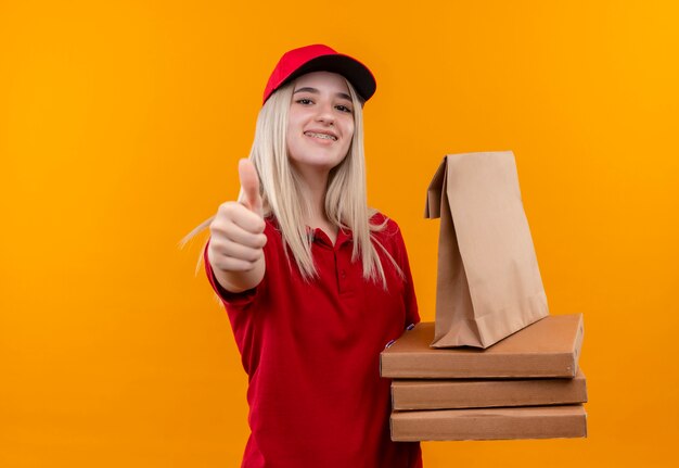 Entrega sonriente joven vestida con camiseta roja y gorra en soporte dental con caja de pizza y bolsillo de papel con el pulgar hacia arriba sobre fondo naranja aislado