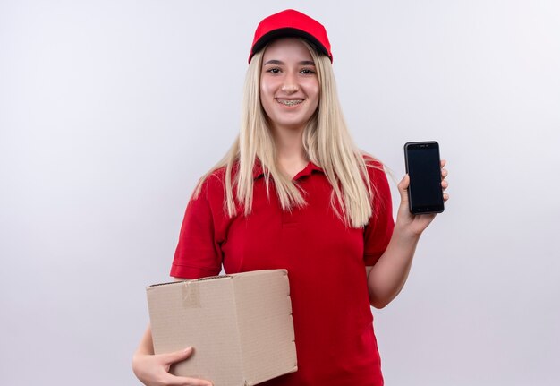 Entrega sonriente joven vestida con camiseta roja y gorra en soporte dental sosteniendo el teléfono y la caja sobre fondo blanco aislado