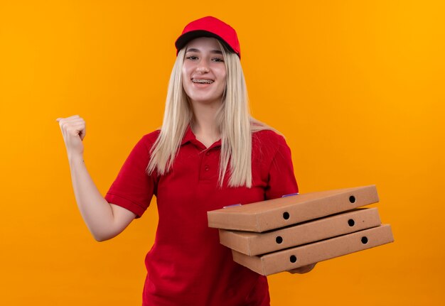 Entrega sonriente joven vestida con camiseta roja y gorra sosteniendo la caja de pizza mostrando sí gesto sobre fondo naranja aislado