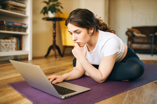 Entrenador de fitness femenino professinal joven atractivo sentado en la estera con computadora portátil con mirada pensativa, grabación de clases de video para entrenamiento en línea. Chica guapa viendo tutorial de yoga en la computadora portátil