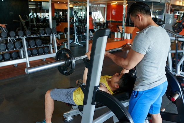 Entrenador personal masculino instruyendo al cliente asiático en press de banca con barra en el gimnasio