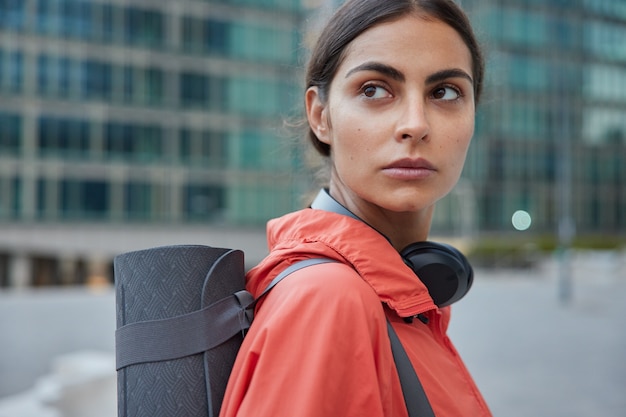 Foto gratuita la entrenadora de yoga femenina espera a que alguien en la calle tiene un descanso de entrenamiento que restaura la fuerza después de que el entrenamiento lleva un estilo de vida saludable se coloca de lado contra la ciudad borrosa