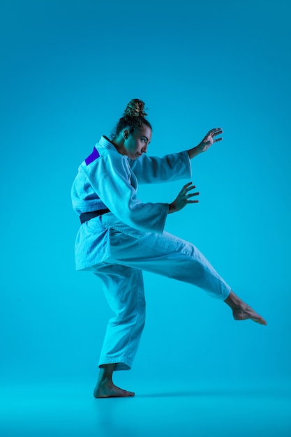 Entrenamiento activo. Judoista profesional femenino en kimono de judo blanco practicando y entrenando aislado sobre fondo azul neón studio.