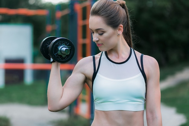 Entrenamiento atlético atractivo de la mujer joven en bíceps con pesa de gimnasia en parque