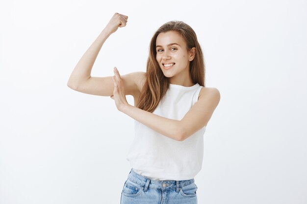 Entrenamiento descarado de chica atractiva en el gimnasio, flexionando bíceps y sonriendo, invitando a comprar membresía, unirse al equipo de fitness