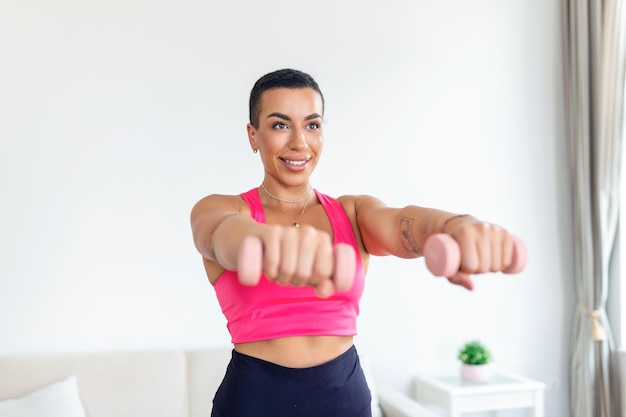 Entrenamiento doméstico con pesas Mujer negra positiva haciendo ejercicios con pesas fortaleciendo su cuerpo en casa Mujer joven sonriente trabajando en sus músculos bíceps manteniéndose saludable