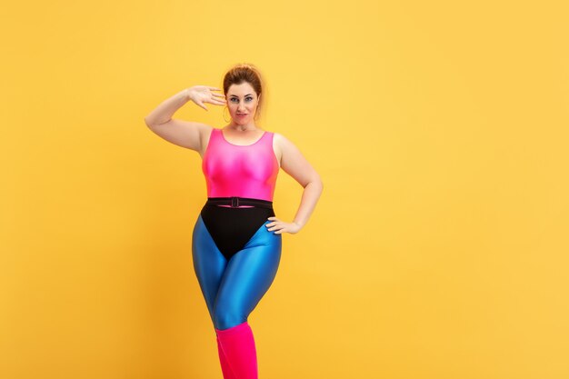 Entrenamiento del modelo femenino caucásico joven del tamaño extra grande en fondo amarillo. Copyspace. Concepto de deporte, estilo de vida saludable, cuerpo positivo, moda, estilo. Mujer elegante posando confiada como superhéroe.