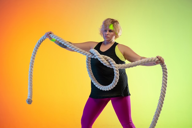 Entrenamiento del modelo femenino caucásico joven del tamaño extra grande en fondo anaranjado degradado en luz de neón. Haciendo ejercicios de entrenamiento con las cuerdas. Concepto de deporte, estilo de vida saludable, cuerpo positivo, igualdad.