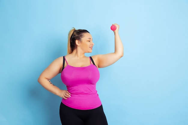 Entrenamiento del modelo femenino caucásico joven del tamaño extra grande en fondo azul. Concepto de deporte, emociones humanas, expresión, estilo de vida saludable, cuerpo positivo, igualdad. Entrenando con las pesas, copyspace.